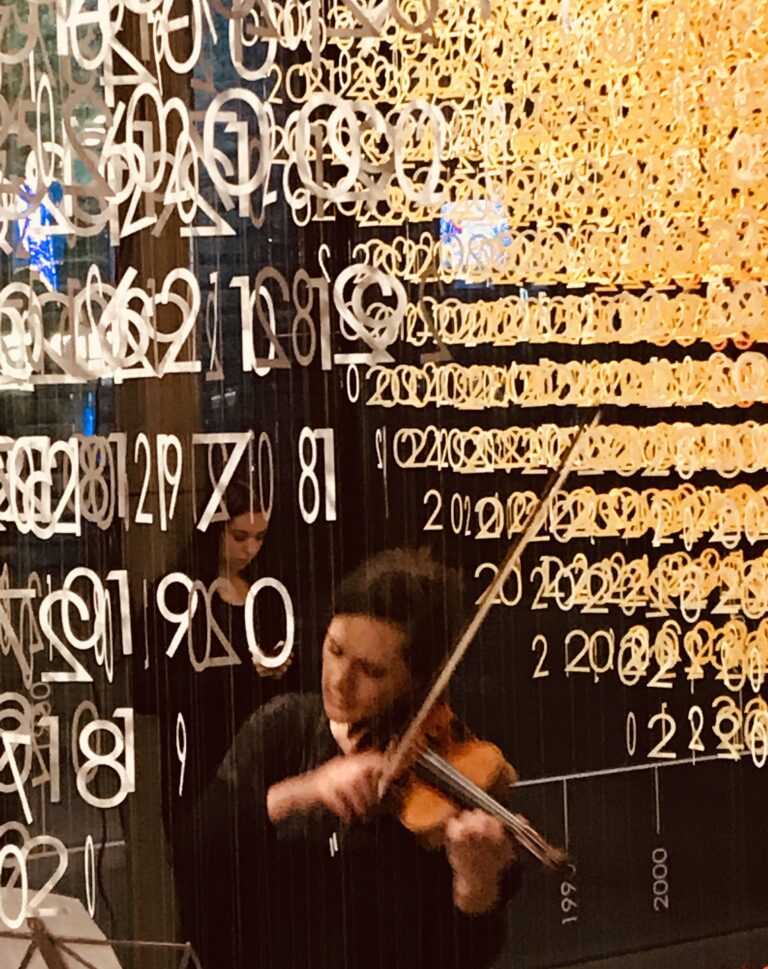 A blurred image of woman playing the violin standing underneath an Emmanuelle Moureaux’s Slices of Time art installation - hundreds of golden numbers suspended in the air.