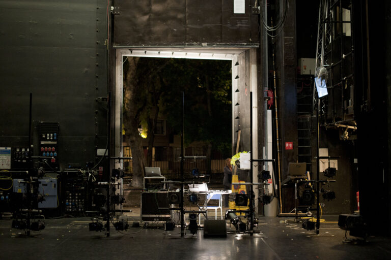 A shot of the side of stage at Sadlers Wells, with the door to outside open so that the street is visible. It is dark outside and we can see a street lamp. The inside of the theatre is dimly lit and we see a collection of technical equipment - a sound desk, a laptop, a chair and a collection of theatre lamps on stands.