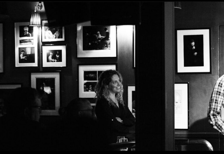 This is a black-and-white photograph of Felicity Hall. Felicity is standing next to a wall in what appears to be a music venue. The wall is covered with several photographs of musicians, mainly piano players, with light coming from out of frame to the right of the photograph and from a lamp on the wall. She is smiling, standing up, has her arms folded, and is looking out to the right of the photograph. Between her and the camera there are two people sat down looking in the same direction as her, there are two glasses on a table, and there is a large pillar.