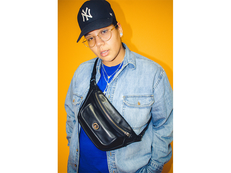 Here we have a photograph of Uniq Being, the musician and teacher who will be leading this workshop for Clod Ensemble. The image features Uniq Being close up to the camera. She is wearing a New York Yankees baseball cap, a blue overshirt, a darker blue undershirt, glasses, necklaces and a black strap over bag. We cannot see her legs. She is standing against a yellow background with her hands in her pockets.